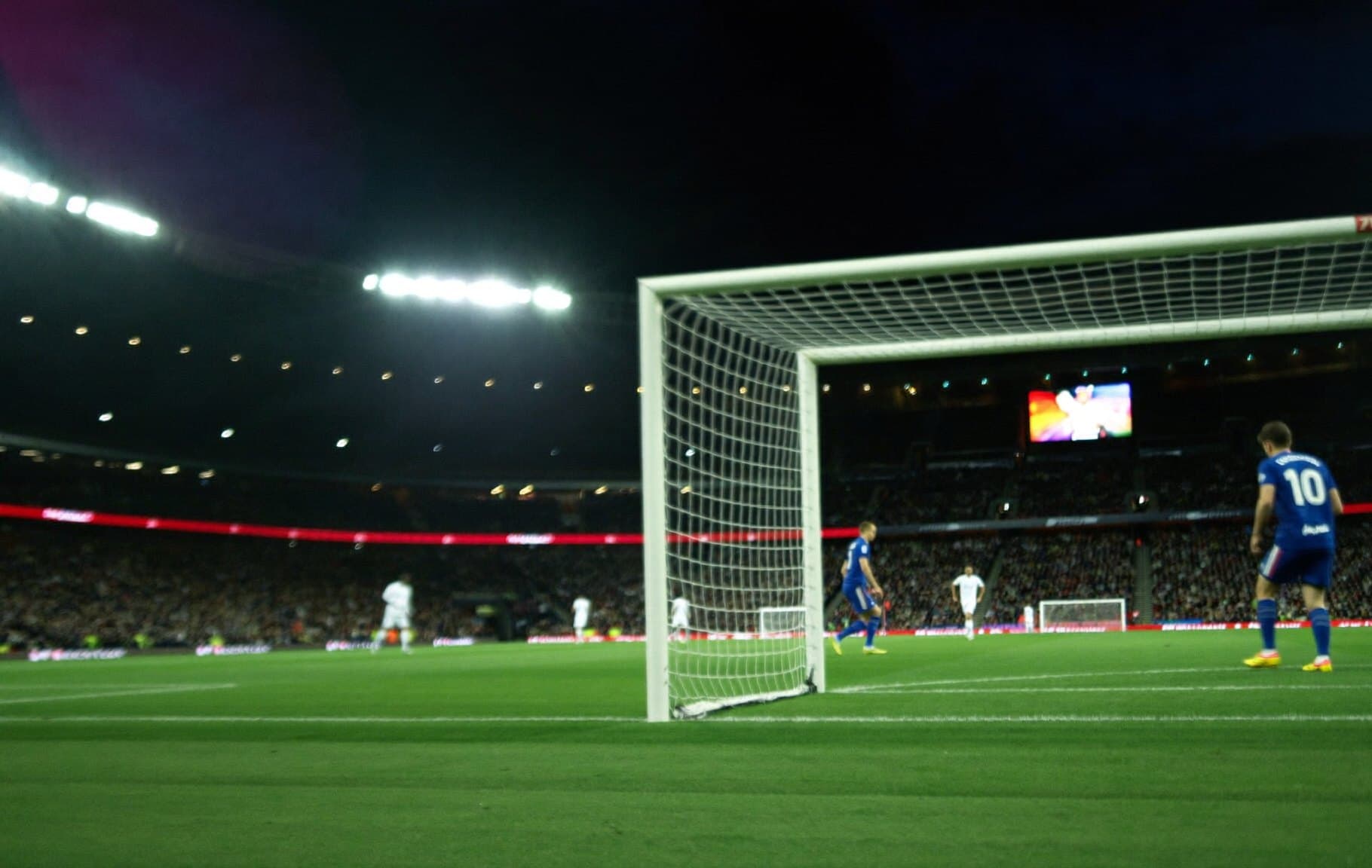 A floodlit football stadium at night seen from behind the goal