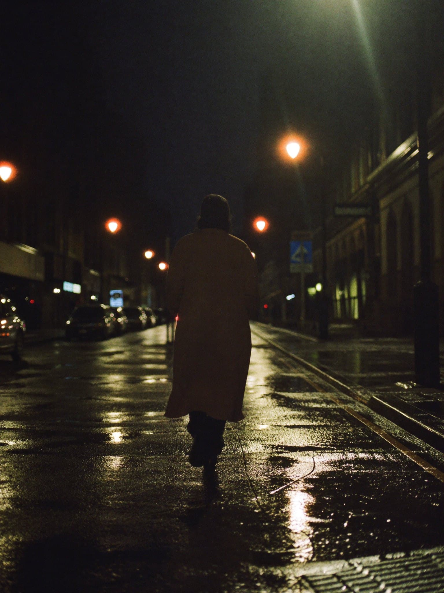 A lone figure walking down a rain soaked city street at night, lit by streetlamps